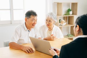 リビングでスーツ姿の女性が、年配の夫婦にパソコンで外壁塗装の説明をしている。安心感のある相談シーン。