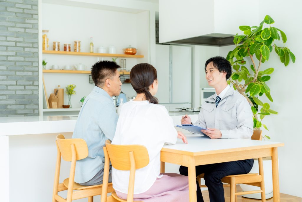 リビングのテーブルで、作業服にネクタイを締めた男性アドバイザーが、見積書を手に夫婦へ説明している様子。外壁塗装の内容を中立の立場で分かりやすく説明しているシーン。