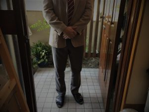 玄関先に立つスーツ姿の男性。顔は映っておらず、外壁塗装などの訪問販売を連想させる構図。